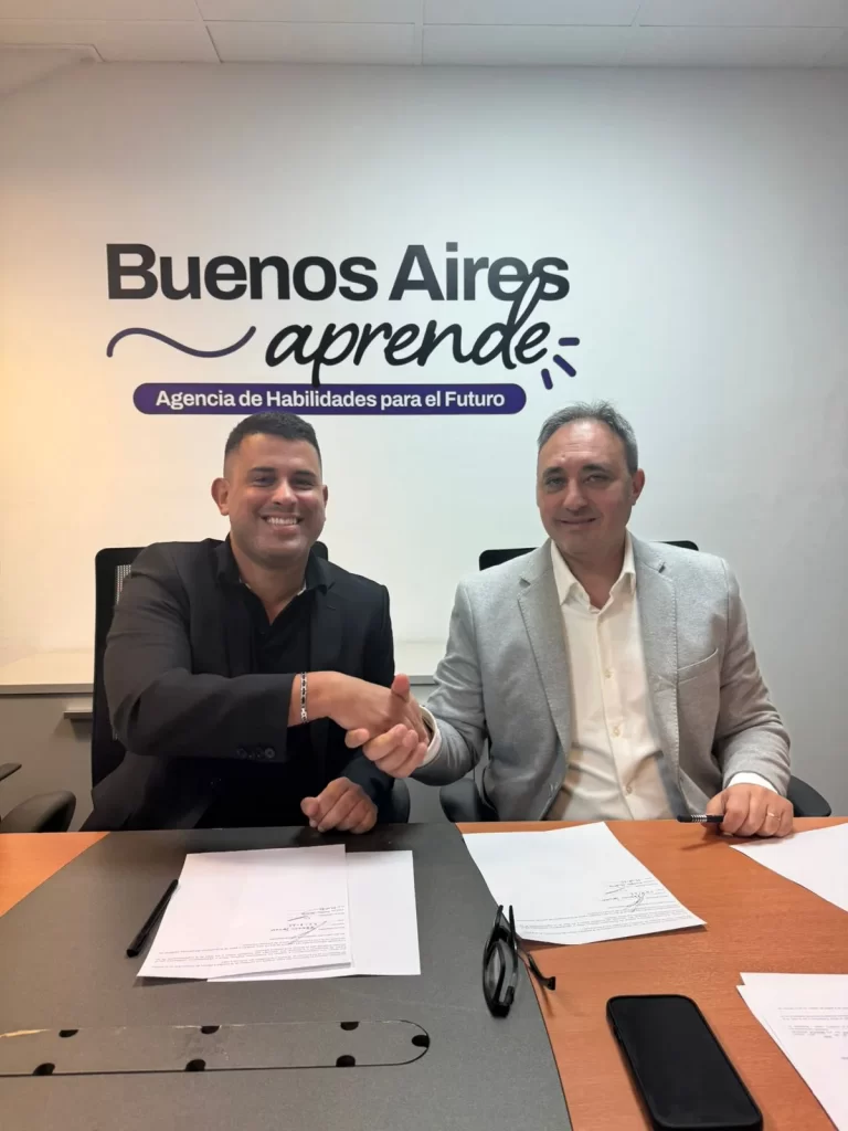 Two men in business attire shake hands at a desk in an office; wall shows the Buenos Aires aprende logo and slogan.es