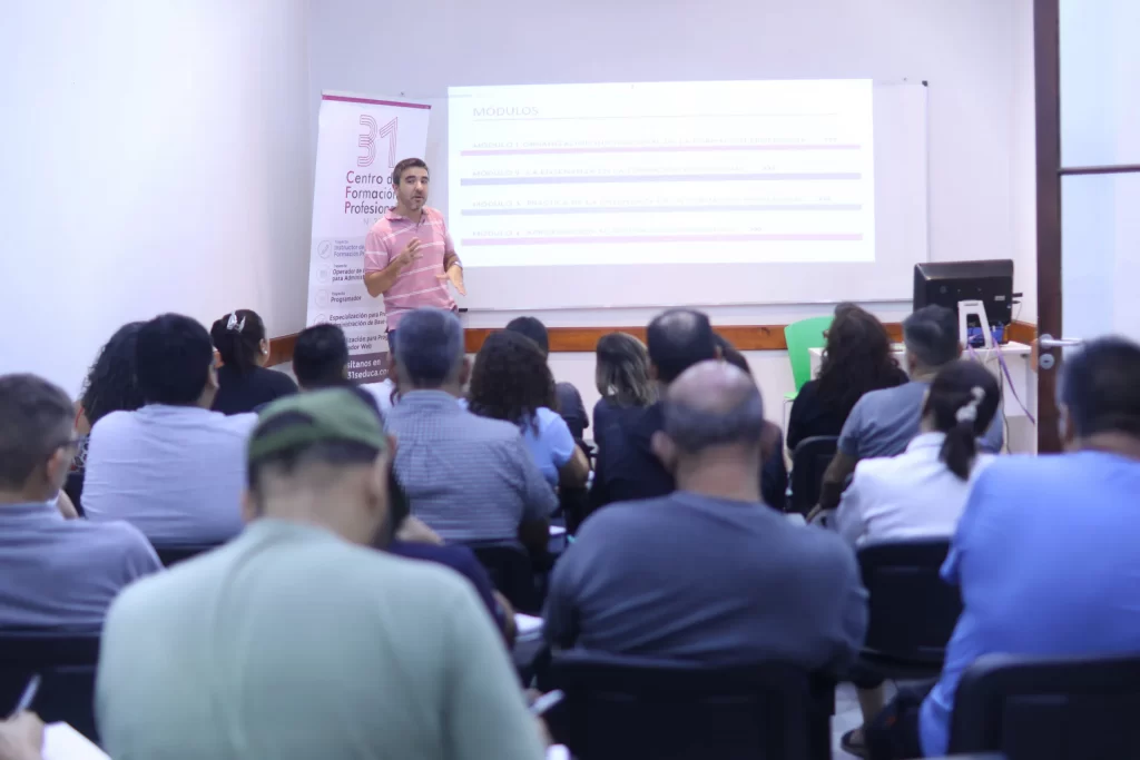 Male presenter in pink polo explains a slideshow to a seated audience in a classroom; projection screen and banner visible at the front.