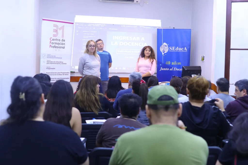 Three presenters stand at the front of a classroom: two women and a man, with banners on either side and a seated audience facing them.
